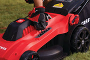 Close-up of a person wearing red and black gloves adjusting the height setting lever on a red CRAFTSMAN corded electric lawn mower, shown on grass.