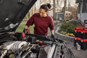 A person wearing gloves and a red sweatshirt is working under the hood of a car outdoors, using a red tool. Nearby, a stack